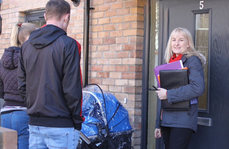 Two tenants wait outside their new home with a pushchair. A housing officer, holding a clipboard, is smiling at the camera whilst stood in front of a front door.