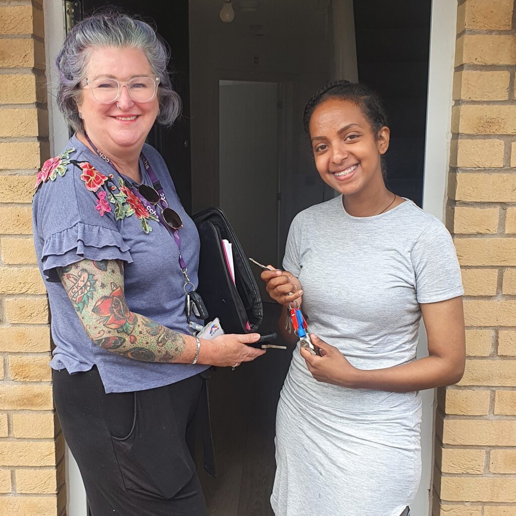 Joni stands next to a tenant at the front door of a house. She is handing over the keys to the new property. Both are smiling at the camera.